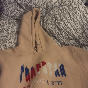 Beige Trapstar hoodie med brodyr - Säljer en beige hoodie från Trapstar med broderad logga i blått, rött och vitt framtill. Tröjan har huva med snörning och en stor magficka. Perfekt för dig som gillar streetwear och oversize. .