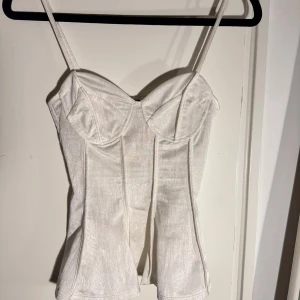 Vit korsett-topp i satin XS - Supersnygg vit korsett-topp i satin med tunna axelband och markerade kupor. Toppen har en lätt glansig finish och sömmar som ger en strukturerad look. Perfekt att styla med jeans eller kjol för en trendig outfit.