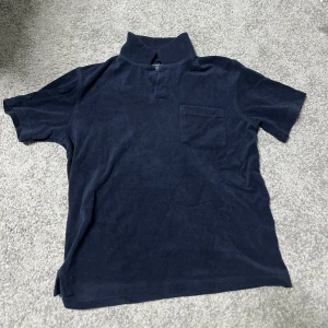 Mörkblå t-shirt från Bondelid - Mörkblå t-shirt från Bondelid med klassisk krage och bröstficka. Tillverkad i mjuk bomull som känns skön mot huden. Perfekt för dig som gillar en stilren och enkel look med en twist. Passformen är normal och ärmarna är korta. Endast använd 1 gång