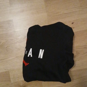 Svart Jordan hoodie med röd logga - Svart hoodie från Jordan med den klassiska röda Jumpman-loggan och vit text på bröstet. Tröjan har huva och en stor magficka framtill. Perfekt för dig som gillar streetwear och sportig stil.