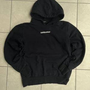 Svart Saint Laurent hoodie med tryck - Svart hoodie från Saint Laurent med vit logga tryckt på bröstet. Klassisk modell med huva och ribbade muddar vid ärmslut och nederkant. Perfekt för dig som gillar stilrena och exklusiva streetwear-plagg.