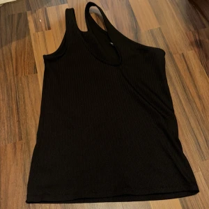 Svart oneshoulder topp Gina Tricot - Säljer en svart ribbad oneshoulder topp från Gina Tricot. Snygg asymmetrisk design med bred axelrem på ena sidan och ärmlös på andra. Perfekt att styla med jeans eller kjol för en trendig look.