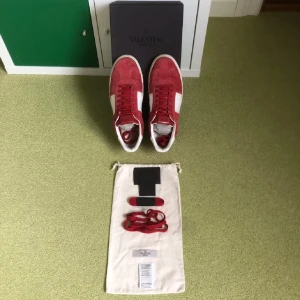 Valentino Flycrews - Säljer ett par Valentino Flycrews sneakers i rött och vitt med snygga detaljer och platt sula. Skorna har snörning, rund tå och är tillverkade i mocka och skinn. Medföljer originalkartong, extra röda skosnören och dusbags. Alla nitar är kvar förutom en som lossnat men den följer med.