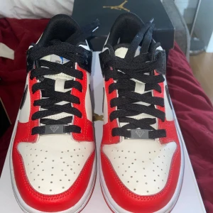 Nike Dunk Low röd/svart/vit skinn - Nike Dunk Low sneakers i skinn med svart swoosh, röda och vita paneler samt svarta skosnören. Klassisk låg modell med perforerad tå och vit mellansula. Röd yttersula och detaljer i svart på häl och snörning. Perfekt för dig som gillar streetwear och snygga färgkombos.