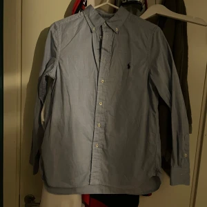 Ljusblå skjorta från Ralph Lauren - Snygg ljusblå skjorta från Ralph Lauren i slim fit-modell. Skjortan har klassisk krage med knappar, lång ärm och den ikoniska broderade loggan på bröstet. Tillverkad i bomull och passar perfekt till en stilren look. I st 14 år aldrig använd 