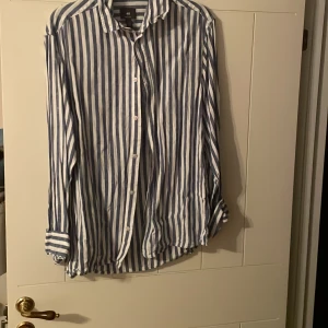 Randig blåvit skjorta från H&M - Säljer en snygg blåvit randig skjorta från H&M i regular fit. Skjortan har långa ärmar, klassisk krage och knäppning framtill. Tillverkad i tunn bomull som känns luftig och skön. Perfekt för dig som gillar en stilren och avslappnad look.