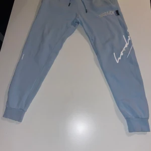 Ljusblå mjukisbyxor från Hoodrich - Snygga ljusblå mjukisbyxor från Hoodrich med vita tryckdetaljer och logga på benet. Byxorna har resår och snörning i midjan samt muddar vid bensluten. Perfekta för en chill och avslappnad stil. Ej andvänd så många gånger (Limited edition)