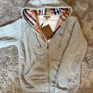 Burberry Zip-Up Hoodie  - Stilren hoodie från Burberry i grått med den klassiska rutiga huvan som exklusiv detalj. En tidlös premium-piece som kombinerar komfort och lyx – perfekt för både vardag och mer uppklädda tillfällen.  ♦️ Helt ny – med originaltag kvar ☑️ ♦️ Exklusiv Burberry-detalj i huvan ☑️  📦 Snabb leverans & trygg affär 