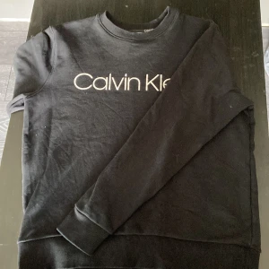 Svart sweatshirt från Calvin Klein - Svart sweatshirt från Calvin Klein med klassisk logga tryckt framtill. Tröjan har rund halsringning, ribbade muddar vid ärmslut och nederkant samt lång ärm. Perfekt för en clean och stilren look.