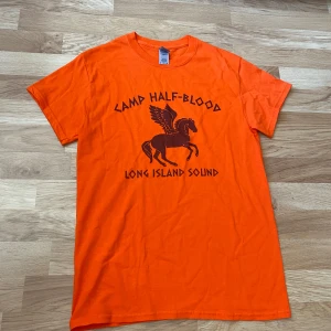 Orange Camp Half-Blood t-shirt - Säljer en orange t-shirt från med Camp Half-Blood-tryck och en Pegasus på bröstet. Klassisk passform med korta ärmar och rund hals. Perfekt för dig som gillar Percy Jackson.