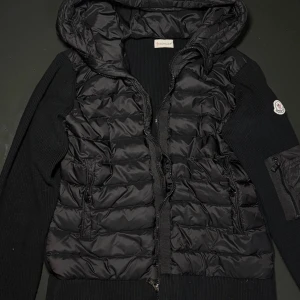 Svart Moncler pufferjacka med huva - Svart pufferjacka från Moncler med stickade ärmar och huva. Jackan har dragkedja framtill, fickor med dragkedja och Moncler-logga på ärmen. Kombinerar quiltad pufferlook med stickade detaljer för en riktigt clean och modern vibe.