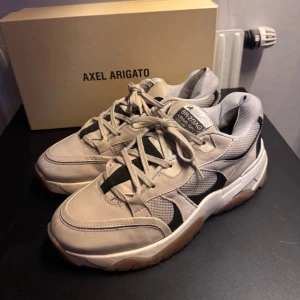 Chunky sneakers från Axel Arigato - Snygga chunky sneakers från Axel Arigato i beige och svart med vit sula och gummidetaljer. Ovandel i skinn och mesh för en cool och sportig look. Klassisk snörning och robust yttersula med Arigato-logga. Perfekta för dig som gillar streetstyle.