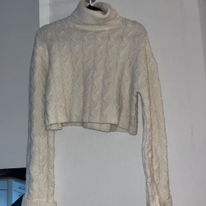 Beige croppad polotröja kabelstickad - Säljer en beige croppad polotröja med kabelstickat mönster. Tröjan har långa ärmar och en hög polokrage, perfekt för kyliga dagar. Den är stickad och har en mjuk känsla, passar dig som gillar en trendig och mysig stil.