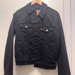 Svart jeansjacka från Levi's - Klassisk svart jeansjacka från Levi's med raka linjer, två bröstfickor med knappar och diskret röd logga. Jackan har långa ärmar, knäppning framtill och är tillverkad i slitstarkt jeanstyg. Perfekt för dig som gillar en tidlös och clean stil.