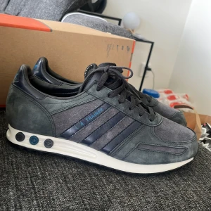 Adidas LA Trainer sneakers grå/blå - Adidas LA Trainer sneakers i mörkgrå mocka och skinn med blå detaljer och klassiska tre ränder på sidan. Snygg retrostil med vit sula och svarta pluggar i hälen. Skorna har snörning och svart yttersula med nitar för extra grepp. Original box ingår 