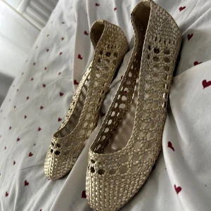Guldiga flätade ballerinaskor - Snygga ballerinaskor i guld med flätad och luftig design. Skorna har rund tå och platt sula, perfekta för dig som vill ha en lätt och somrig look. Materialet är syntet och de har ett elegant, glittrigt intryck som sticker ut.