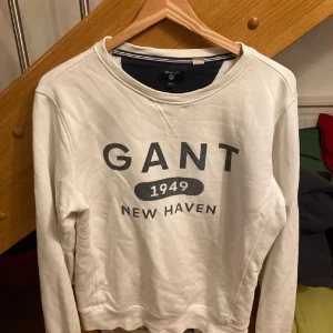 Vit sweatshirt från GANT med tryck - Vit sweatshirt från GANT med svart tryck framtill där det står 'GANT 1949 NEW HAVEN'. Tröjan har rund halsringning, långa ärmar och är tillverkad i mjuk bomull. Perfekt för en chill och stilren look.