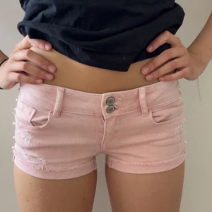 Ljusrosa jeansshorts med pärldetaljer - Säljer mina snyggaste ljusrosa jeansshorts. Shortsen har pärldetaljer längs sidorna och dubbla knappar framtill. Perfekta för sommaren💖💖Passar som XS