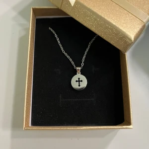 Silverhalsband med korsberlock - Halsband i silverfärg med en rund berlock som har ett utskuret kors i mitten. Kedjan är tunn. 