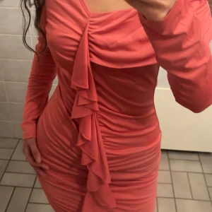 Korallrosa omlottklänning med volang - Supersnygg korallrosa kortklänning med långa ärmar och omlott-detalj framtill. Klänningen har volang längs framsidan och är figurnära med rynkade partier som ger en cool och trendig look. Perfekt för dig som vill sticka ut med färg och form! Passar m också. Aldrig använd 