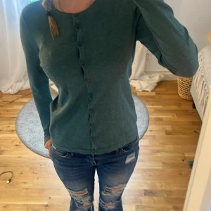 Grön kofta med knappar - Mysig grön kofta med rund hals och knäppning hela vägen framtill. Långärmad och i mjukt material som passar perfekt till jeans eller kjol. Enkel och stilren design som funkar året runt.