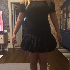 Svart offshoulder kortklänning - Supersöt svart kortklänning med offshoulder och puffig volangkjol. Smockad överdel som sitter tight och markerar midjan. Kjolen har en ballongliknande form som ger en cool och lekfull vibe. Perfekt för dig som vill sticka ut med en trendig look.