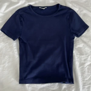 Marinblå basic topp från Gina Tricot - Enkel och stilren marinblå topp från Gina Tricot, basically basics-kollektionen. T-shirten har rund halsringning och korta ärmar, perfekt för en clean och avslappnad look. Gjord i mjukt material som känns skönt mot huden.