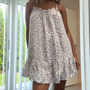 Blommig kort sommarklänning med volang - Supersöt kort klänning med tunna axelband och volang nertill. Ljus botten med små blommor i blått och rosa. Lös passform, perfekt för varma dagar. Mjuk och skön i bomull.