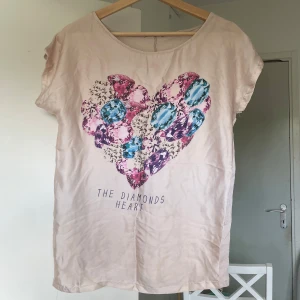 Beige t-shirt med hjärtmotiv - Snygg beige t-shirt med korta ärmar och stort hjärtmotiv av färgglada diamanter i rosa, blått och lila på bröstet. Mjuk och skön, perfekt för en chill look.