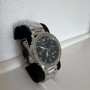 Emporio Armani silverklocka - Stilren klocka från Emporio Armani med silverfärgad länk i rostfritt stål och mörkblå urtavla. Klockan har kronograf, tachymeter och datumvisning. Snygg och modern design med tydliga detaljer och logga upptill på urtavlan.