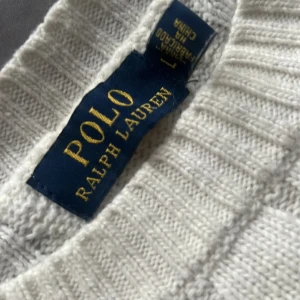 Vit kabelstickad tröja Polo Ralph Lauren - Vit kabelstickad tröja från Polo Ralph Lauren med klassisk rund halsringning och broderad logga på bröstet. Tröjan är i ullblandning och har ribbade muddar vid ärmslut och nederkant. Perfekt för dig som gillar en clean och tidlös stil.