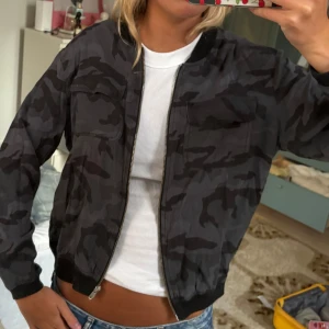 Svart camo bomberjacka med dragkedja - Säljer en svart bomberjacka med mörkgrått camouflage-mönster. Jackan har två bröstfickor med lock, ribbade muddar och hel dragkedja framtill. 