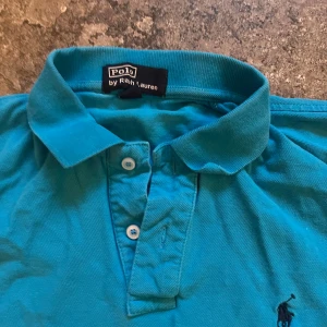 Ralph Lauren Pike Blå - Snygg turkos pikétröja från Polo Ralph Lauren med klassisk krage, två knappar och den ikoniska broderade loggan på bröstet. Tröjan är i bomull och har korta ärmar, perfekt för en chill och sportig stil. strlk l passae m