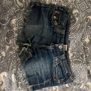 True Religion jeansshorts med fransar - Snygga blå jeansshorts från True Religion med slitna kanter och fransar. Klassiska femficksdetaljer, synliga sömmar och ikoniska bakfickor med lock och knapp. Perfekta för sommaren och ger en cool, avslappnad vibe.