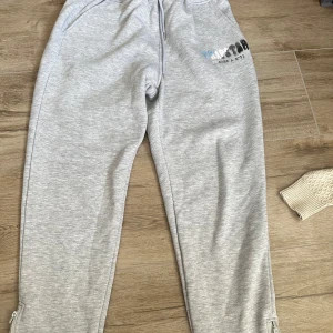 Grå mjukisbyxor från Trapstar L - Snygga grå mjukisbyxor från Trapstar med broderad logga i blått, vitt och svart på ena benet. Byxorna har dragsko i midjan, muddar vid bensluten och dragkedjor vid anklarna. Perfekta för chill eller streetwear.