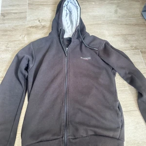 Två McKenzie hoodies, grå & svart - Säljer två hoodies från McKenzie. En är ljusgrå i storlek S med magficka och dragsko i huvan, den andra är svart i storlek M med dragkedja och grått foder i huvan. Båda har tryckt logga på bröstet och är i mjuk bomullsblandning. Perfekta för chill dagar.