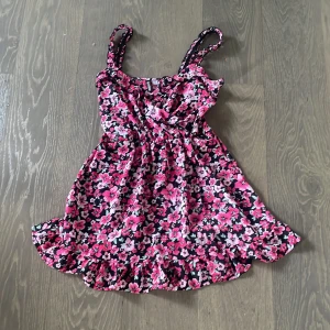 Blommig omlott kortklänning från H&M - Supersöt omlott kortklänning med volangkant och smockade axelband från H&M. Klänningen har ett färgstarkt blommönster i rosa, vitt och svart. Perfekt för varma dagar med sin lätta och luftiga känsla.