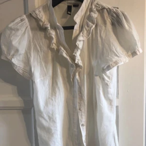 Vit blus med volang och puffärm - Söt vit blus med korta puffärmar och volang längs knäppningen. Blusen har knappar framtill och en feminin, romantisk stil. Perfekt för dig som gillar detaljer och en luftig känsla.