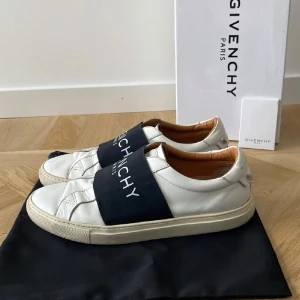 Givenchy vita sneakers med svart band - Snygga vita sneakers från Givenchy med bred svart resår över vristen där loggan står tryckt i vitt. Skorna är i skinn med beige innersida och klassisk platt sula. Perfekta för dig som vill ha en clean och lyxig look med streetkänsla.