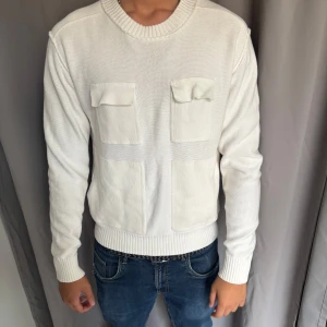 Vit stickad tröja Tiger of Sweden - Vit stickad tröja från Tiger of Sweden i storlek M. Tröjan har två stora bröstfickor med lock och två nedre fickor framtill. Perfekt för en clean och stilren look.