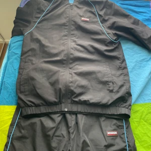 Mckenzie Tracksuit - Svart tracksuit från McKenzie med elastisk midja och blå piping längs sidorna. Byxorna har meshfoder för extra komfort och sportig känsla. Perfekta för chill eller träning, med klassisk McKenzie-logga framtill. har aldrig använt den, pris kan diskuteras ‼️ om man vill köpa separat koftan - 340kr byxan - 220kr