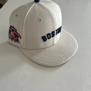 Vit Boston Red Sox keps från New Era - Snygg vit 59FIFTY keps från New Era med broderad BOSTON-text i mörkblått framtill och 1903 World Series patch på sidan. Klassisk flat peak med grön undersida och MLB-logga bak. Tillverkad i ullblandning, perfekt för dig som gillar baseball och streetstyle. Den äe helt ny aldrig använd