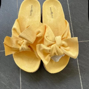 Gula sandaler med stor rosett från Marquitiz - Snygga gula sandaler från Marquitiz med breda band i mocka och stora rosetter framtill. Platt sula med korkdetalj på sidan och vågigt mönster undertill. Perfekta för en somrig look och riktigt bekväma att glida runt i.