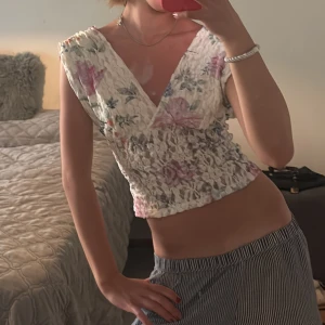 Blommig spetsblus från Gina Tricot XS - Supersöt blommig blus från Gina Tricot i spets med v-ringning och croppad passform. Toppen är vit med rosa och blå blommor, har breda axelband och smockad midja. Perfekt för sommarens alla tillfällen när du vill känna dig extra fin.