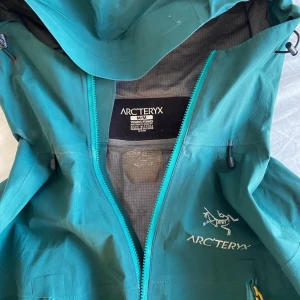 Turkos Arc'teryx Gore-Tex vindjacka dam M - Arc'teryx vindjacka i turkos med GORE-TEX Pro-material, perfekt för blåsiga och regniga dagar. Jackan har huva, dragkedjor i kontrasterande färg och snygga detaljer som gul zip-puller. Logga på bröstet och vattentäta fickor.