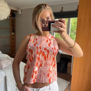 Blommig ärmlös linnetopp i orange och vitt - Säljer en ärmlös linnetopp med blommigt mönster i orange och vitt. Toppen har rund halsringning och små pärldetaljer vid axlarna