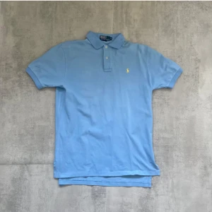 Ljusblå pikétröja från Ralph Lauren - Klassisk ljusblå pikétröja från Polo Ralph Lauren med gul broderad logga på bröstet. Tröjan har korta ärmar, krage och två knappar framtill. Tillverkad i bomull för en skön och luftig känsla. Perfekt för en stilren och avslappnad look.