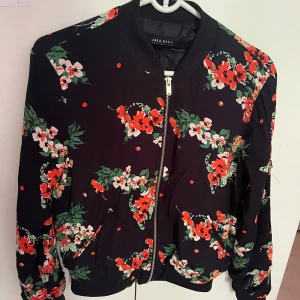 Blommig bomberjacka från Zara Basic - Svart bomberjacka från Zara Basic med färgglatt blommönster i rött, vitt och grönt. Jackan har dragkedja framtill, ribbade muddar och klassisk bomberkrage. Perfekt för dig som vill sticka ut med en cool och trendig look.