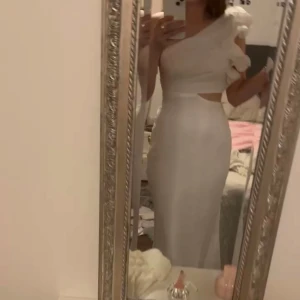 Vit långklänning med volang och cut out - Säljer en elegant vit långklänning med oneshoulder och volangdetalj på axeln. Klänningen har en snygg cut out vid midjan och figurnära passform. Perfekt för dig som vill sticka ut med en stilren och modern look.