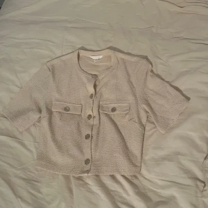 Beige croppad skjorta med knappar - Snygg croppad skjorta i beige med korta ärmar och markerad struktur från h&m.  Skjortan har två bröstfickor med knappar och raka kanter. Perfekt för dig som gillar en stilren och trendig look.